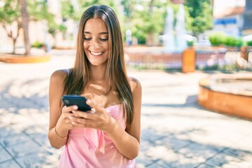 Wall Mural - Young hispanic woman smiling happy using smartphone walking at the park