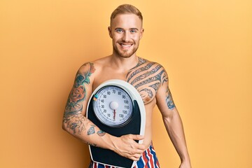 Wall Mural - Young caucasian man holding weight machine to balance weight loss looking positive and happy standing and smiling with a confident smile showing teeth