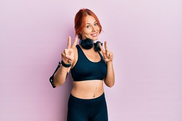 Sticker - Young beautiful redhead woman wearing gym clothes and using headphones smiling looking to the camera showing fingers doing victory sign. number two.