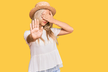 Poster - Young blonde girl wearing summer hat covering eyes with hands and doing stop gesture with sad and fear expression. embarrassed and negative concept.