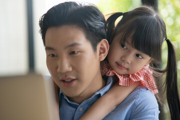 Wall Mural - Asian father on blue shirt working from home with his little cute girl daughter. Asian kid hug his dad from behind while her father is working on a laptop.