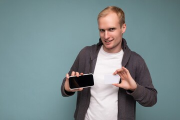 Handsome man wearing everyday clothes isolated on background wall holding and using phone and credit card making payment looking at camera
