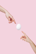 Two Hands Touching Flying Egg On Pastel Pink Background. Creation Of New Life. Minimal Easter Art Concept.