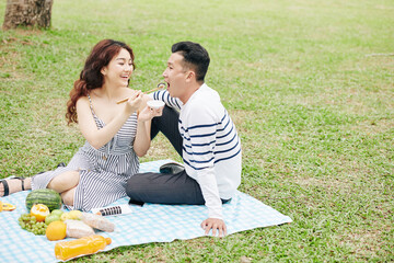 Wall Mural - Pretty smiling young Vietnamese woman feeding boyfriend with delicious suchi when having picninc in park