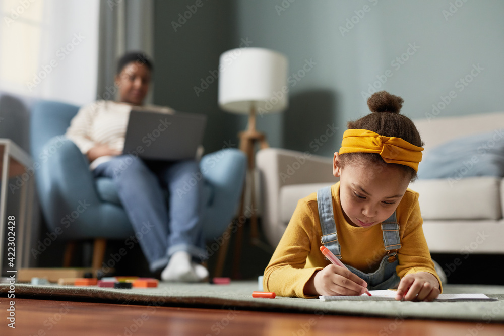 Front view portrait of cute African-American girl drawing while lying on floor in home interior with mother working in background, copy space - obrazy, fototapety, plakaty 