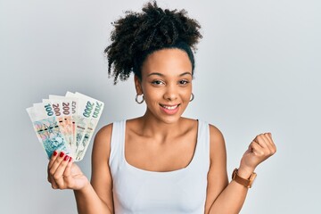 Poster - Young african american girl holding czech koruna banknotes screaming proud, celebrating victory and success very excited with raised arm