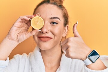 Sticker - Young blonde woman wearing bathrobe holding lemon over eye smiling happy and positive, thumb up doing excellent and approval sign