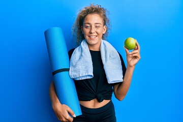 Poster - Beautiful caucasian teenager girl holding yoga mat and green apple winking looking at the camera with sexy expression, cheerful and happy face.