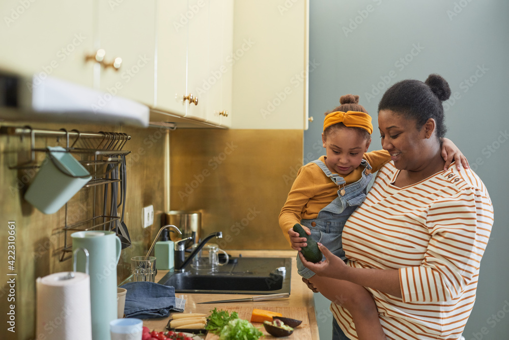 Waist up portrait of happy African-American mother holding daughter while cooking together in kitchen interior, copy space - obrazy, fototapety, plakaty 