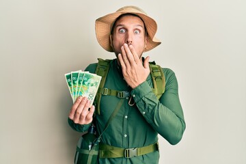 Poster - Handsome man with beard wearing explorer hat holding 50 shekels covering mouth with hand, shocked and afraid for mistake. surprised expression