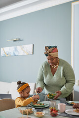 Vertical portrait of African-American family enjoying dinner together in cozy home interior, focus on grandmother serving food to cute little girl, copy space