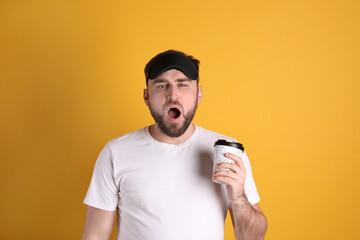 Wall Mural - Tired young man with sleep mask and cup of coffee yawning on yellow background