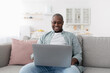 © Prostock-studio - Free time. Happy middle-aged black man using laptop while resting on sofa at home