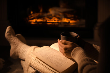 Poster - Woman with book and cup near fireplace indoors, closeup. Cozy atmosphere