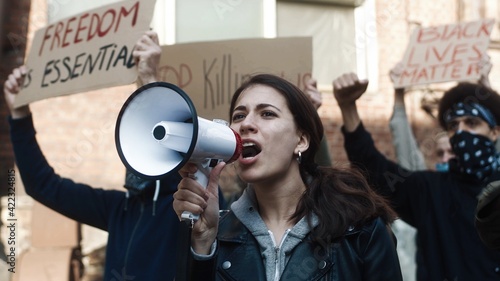 Pretty young Caucasian girl screaming in megaphone while standing among people at protest against racism and police brutality. Mixed-races male and female protesters.