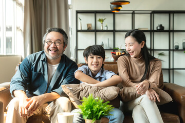 Wall Mural - Happy cheerful Asian family parent with child boy son watching tv together spending holiday weekend time in quarantine on sofa in living room. family and home concept.