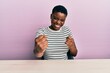 © Krakenimages.com - Young african american woman wearing casual clothes sitting on the table very happy and excited doing winner gesture with arms raised, smiling and screaming for success. celebration concept.