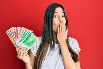 Poster - Young brunette woman holding norwegian krone banknotes covering mouth with hand, shocked and afraid for mistake. surprised expression