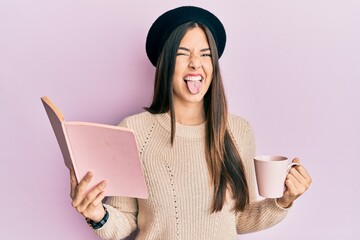 Poster - Young brunette woman reading a book and drinking a cup of coffee sticking tongue out happy with funny expression.