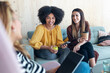 © nenetus - Smart entrepreneur multiethnic women talking while taking a break and showing photos with smart phone sitting on couch at coworking place.