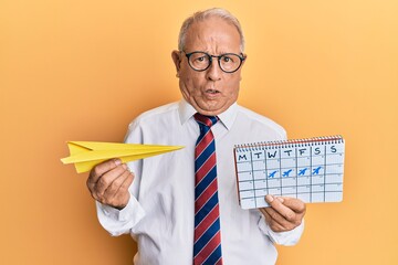 Wall Mural - Senior caucasian man going on business trip holding paper airplane in shock face, looking skeptical and sarcastic, surprised with open mouth