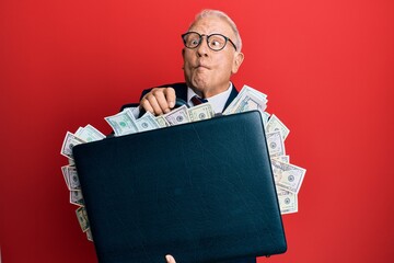 Wall Mural - Senior caucasian man holding briefcase full of dollars making fish face with mouth and squinting eyes, crazy and comical.