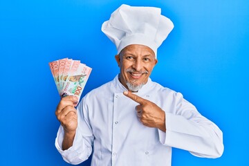 Middle age grey-haired man wearing professional cook uniform holding new zealand dollars banknotes smiling happy pointing with hand and finger