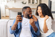 © Vadim Pastuh - Excited and overjoyed African-American young couple sits on the couch at home and holds smartphone, a man and woman show victory gesture, celebrating good news or win, received money on account