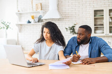Concentrated African-American Couple Sits At The Kitchen Table With A Laptop At Home, Calculating Finance, Checking Bank Account Balance, A Biracial Woman Looks Through Papers, A Man Takes A Notes