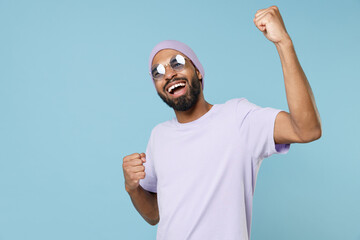 Wall Mural - Young smiling happy cool unshaven black dark-skinned african man 20s in violet t-shirt purple hat glasses do winner gesture clench fist celebrating isolated on pastel blue background studio portrait