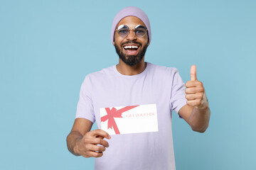 Wall Mural - Young happy smiling satisfied unshaven student black african man in violet t-shirt hat glasses hold gift voucher flyer mock up show thumb up gesture isolated on pastel blue background studio portrait.