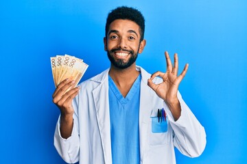 Wall Mural - Handsome hispanic man with beard wearing medical uniform holding 500 norwegian krone doing ok sign with fingers, smiling friendly gesturing excellent symbol