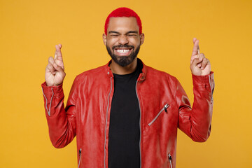 Canvas Print - Young african man 20s with funky trendy pink hairdo in red leather jacket wait for special moment keep fingers crossed making wish, eyes closed isolated on yellow orange background studio portrait.