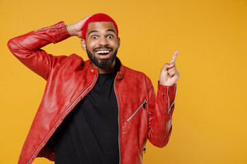 Wall Mural - Young african dark-skinned shocked man with funky trendy pink hairdo in red leather jacket point index finger overhead on workspace area hold face isolated on yellow orange background studio portrait.