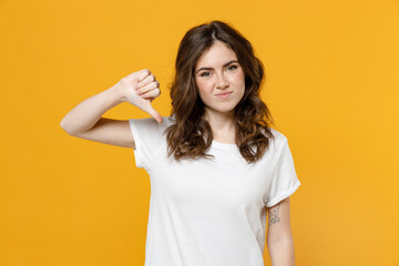 Young sad disappointed displeased caucasian student woman 20s in white basic casual blank print design t-shirt show thumb down dislike gesture isolated on yellow orange background studio portrait.