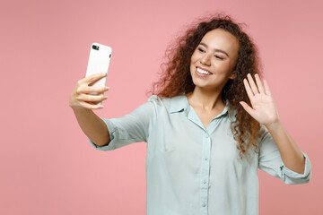 Wall Mural - Young black african curly friendly woman 20s wear casual blue shirt doing selfie shot on mobile phone talking by video call waving hand hello isolated on pastel pink color background studio portrait.