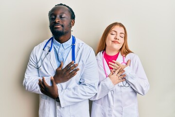 Canvas Print - Young interracial couple wearing doctor uniform and stethoscope smiling with hands on chest with closed eyes and grateful gesture on face. health concept.