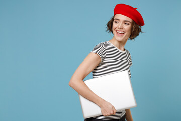 Wall Mural - Side profile view young freelancer woman 20s with short hairdo wear french beret red hat striped t-shirt hold closed laptop pc computer look aside isolated on pastel blue background studio portrait.