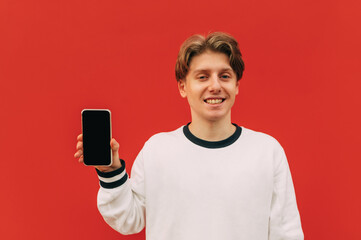 Wall Mural - Happy guy in standing on a red background with a smile on his face and showing a smartphone with a black screen to the camera on a red wall background.