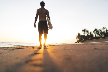 Back view of man holding professional surfboard enjoying sunset promenade during weekend time for hobby practice, sportive male surfer walking during summer vacations at Punalu'u Beach in Hawaii