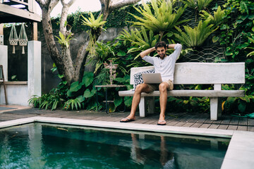 Wall Mural - Excited young man entertaining with laptop near swimming pool in tropical resort