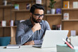 © insta_photos - Young serious indian professional business man, focused ethnic male student wearing glasses working on laptop, remote studying using computer looking at screen watching seminar webinar at home office.