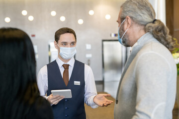 Canvas Print - Young male receptionist in protective mask and formalwear using touchpad while talking to mature businessman in the lounge