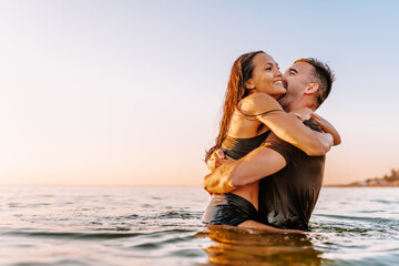 Happy young fit couple in sea or ocean hug each other with love at summer sunset. Romantic mood, tenderness, relationship, vacation concept.