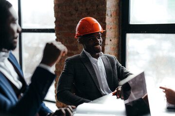 Wall Mural - An African American man in an orange construction helmet provides the blueprints for the construction of a building to investors. An engineer sits in an office at a table near the panoramic windows