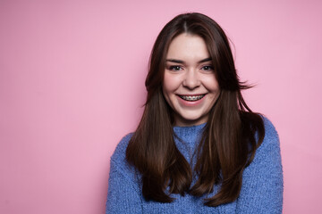 Portrait of an attractive brunette girl in braces, in a blue sweater smiling on a pink background. Dentistry concept, beautiful smile