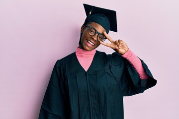 Wall Mural - Young african american girl wearing graduation cap and ceremony robe doing peace symbol with fingers over face, smiling cheerful showing victory