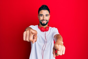 Canvas Print - Young man with beard listening to music using headphones pointing to you and the camera with fingers, smiling positive and cheerful