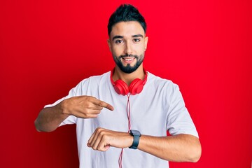 Wall Mural - Young man with beard listening to music using headphones in hurry pointing to watch time, impatience, upset and angry for deadline delay