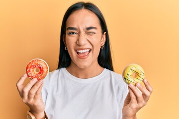 Poster - Young asian woman holding tasty colorful doughnuts winking looking at the camera with sexy expression, cheerful and happy face.